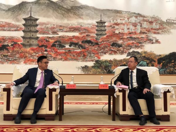 Two men in suits seated in armchairs, facing each other across a small table, in front of landscape art.